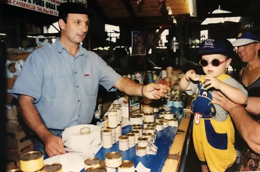 Vente de Foie Gras : Marché et Enfant Stylé Vendeur souriant présentant des conserves de foie gras à un bambin avec lunettes de soleil et salopette de dinosaure. Texte: "FOIES GRAS" et "05 53".