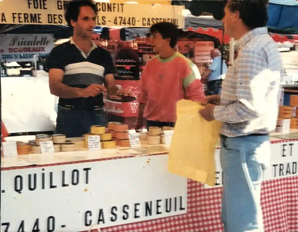 Marché : Vente de foie gras Quillot à Casseneuil Vendeur de foie gras et confit 'Quillot - Casseneuil' présentant des conserves à des clients sur un marché.