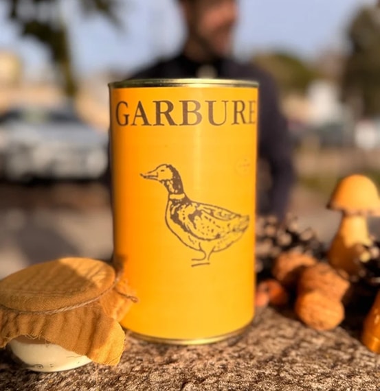 Boîte de conserve jaune "GARBURE" avec l'illustration d'un canard, posée à l'extérieur avec un bocal et des décorations de champignons.