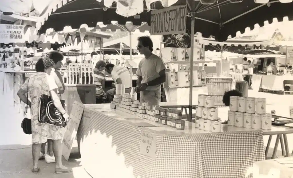 Marché de produits fermiers : Vente de conserves et sandwichs Vendeur aux lunettes de soleil sur un stand de marché proposant des produits fermiers. Panneau : "PRODUITS FERMIERS". Prix affiché : "Sandwich au Ria 6".