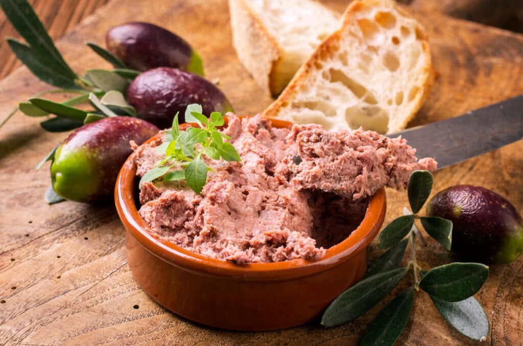 Pâté rustique, olives et pain frais sur bois Pâté de campagne rustique dans un ramequin en terre cuite, servi avec des olives noires et du pain à la coupe sur planche de bois.