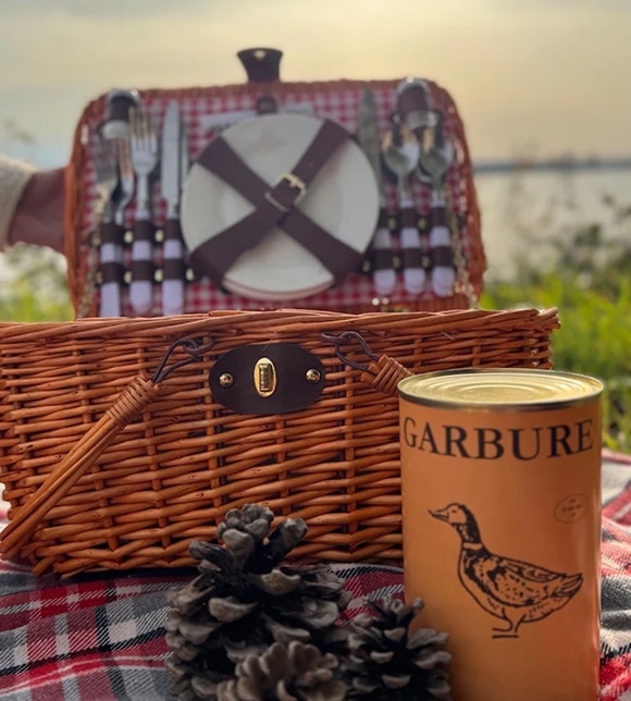Pique-nique champêtre : Garbure et panier en osier Pique-nique au coucher du soleil avec un panier en osier, une conserve de GARBURE illustrée d'un canard, et des pommes de pin sur une couverture à carreaux.